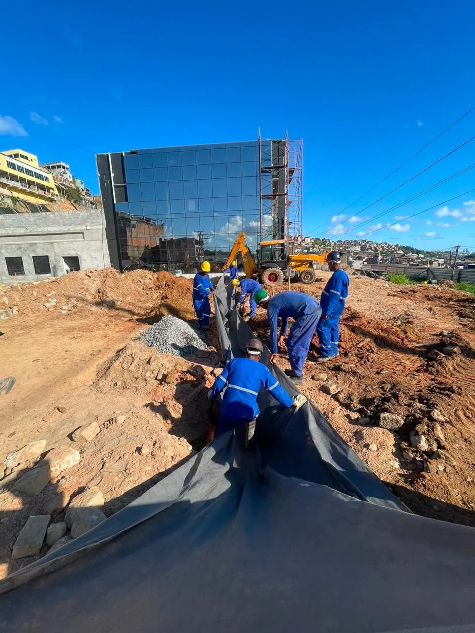 Equipe da Roble Serviços instalando manta geotêxtil em obra de drenagem, com retroescavadeira ao fundo